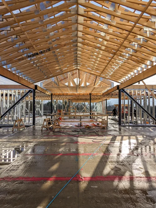 Construction site of a building with a wooden roof structure, metal framing, and a concrete floor that is wet and partially coated with a liquid substance. There are tools, wooden sawhorses, and construction workers present.